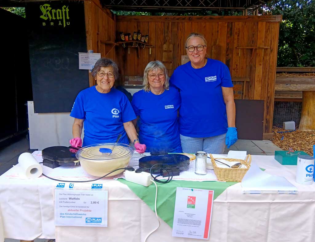 Three smiling women in matching blue shirts and gloves stand behind a table with cooking equipment, food items, and signs, at an outdoor wooden booth.